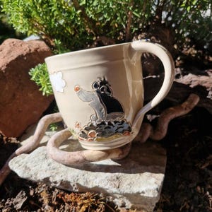 Könnte beinhalten: Ein cremefarbener Keramikbecher mit einer erhabenen, handbemalten Illustration eines schwarzen und grauen Maulwurfs, der aus einem Erdhügel hervorkommt. Das Design zeigt eine kleine weiße Wolke und einen geschwungenen Henkel an der rechten Seite.
