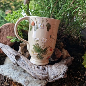 Op de afbeelding: Een crèmekleurige keramische mok met reliëf en geschilderde botanische details, waaronder groene bladeren, witte bloemen, kleine rode aardbeien en een libel. De mok heeft een gebogen handvat en een voetstuk met een gladde afwerking.