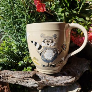 Könnte beinhalten: Beigefarbene Keramiktasse mit einem Cartoon-Waschbären-Design. Der Waschbär ist in Schwarz, Grau und Weiß dargestellt und hält ein kleines Objekt. Die Tasse hat einen großen Henkel. Der Hintergrund zeigt Grün und rote Blumen.