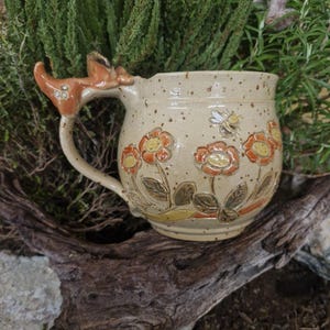 Könnte beinhalten: Eine beige Keramiktasse mit einem hirschförmigen Griff. Die Tasse ist mit orangefarbenen und gelben Blumen, grünen Blättern und einer kleinen Biene verziert. Die Tasse steht auf einem Stück Treibholz.