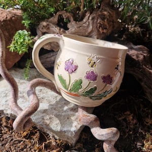 Könnte beinhalten: Ein cremefarbener Keramikbecher mit geprägten und bemalten Details von violetten Blumen mit grünen Blättern sowie kleinen gelben und schwarzen Bienen. Der Becher hat einen abgerundeten Körper und einen geschwungenen Henkel mit glatter Oberfläche.