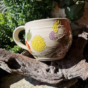 Può includere: Tazza in ceramica color crema con un motivo floreale. La tazza presenta fiori gialli, viola e arancioni in rilievo con foglie verdi. La tazza ha un manico curvo ed è ambientata su uno sfondo naturale.