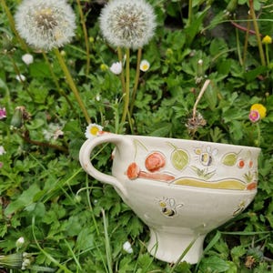 Op de afbeelding: Een crèmekleurige keramische theekop met een voetstuk en een gebogen handvat. De beker is versierd met geschilderde bijen, bladeren en bessen. De theekop staat in een bed van groen gras met paardenbloemen en kleine witte en gele bloemen.
