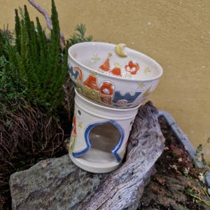 Könnte beinhalten: Ein cremefarbener Keramik-Duftlampen-Wärmer mit dekorativem Design. Die obere Schale zeigt farbenfrohe Illustrationen von Häusern, Sternen und einem Mond. Der Sockel hat eine bogenförmige Öffnung, die blau umrandet ist. Der Wärmer steht auf einem Stein.