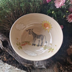 Könnte beinhalten: Eine beige Keramikschale mit handgemaltem Esel-, Sonnen- und Wolken-Design. Die Schale hat einen breiten Rand und eine geringe Tiefe. Der Esel ist grau, und die Sonne ist gelb und orange. Die Schale ist mit Blumen und Wolken verziert.