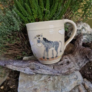 Könnte beinhalten: Beige Keramikbecher mit einer skurrilen Esel-Illustration. Der Esel ist grau mit schwarzen Akzenten und steht auf einem braunen und orangefarbenen Sockel. Weiße Wolken sind um den Esel herum verstreut. Der Becher hat einen gebogenen Henkel und ist von Grün umgeben.