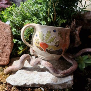Könnte beinhalten: Eine beige Keramiktasse mit Henkel, verziert mit farbenfrohen botanischen Designs. Die Tasse hat orange, grüne und blaue Akzente. Sie steht auf einer Steinoberfläche mit Grünzeug im Hintergrund, was eine natürliche Ästhetik erzeugt.