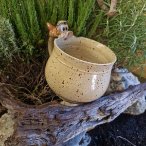Puede incluir: Una taza de cerámica beige moteada con un asa en forma de dragón. La taza tiene forma redonda y una base pequeña. El asa del dragón es marrón y beige. La taza está colocada sobre un trozo de madera flotante, con plantas verdes al fondo.