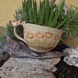 Puede incluir: Una taza de cerámica blanca con asa negra y el texto "But First, Coffee" en negrita negra. La taza se encuentra sobre un fondo borroso, sugiriendo un ambiente acogedor.