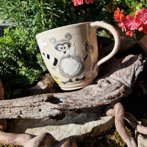 Puede incluir: Taza de cerámica color crema con un diseño de mapache de dibujos animados. El mapache tiene una cola a rayas blancas y negras y sostiene un letrero en forma de corazón. La taza tiene un asa curva. Rodeada de vegetación y flores rojas.