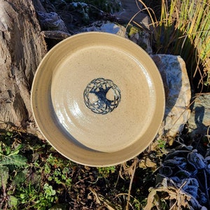 Op de afbeelding: Een beige keramische kom met een blauw levensboomontwerp in het midden. De kom staat op een houten oppervlak.