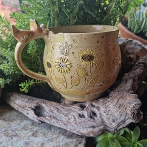 Puede incluir: Una taza de cerámica beige moteada con diseños en relieve de girasoles y una abeja en el exterior. El asa tiene forma de pequeño animal con orejas y patas visibles. La taza presenta un cuerpo redondeado y un acabado esmaltado uniforme.
