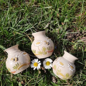 Könnte beinhalten: Drei kleine, beige Keramikvasen mit Blumen- und Bienenmotiven, angeordnet auf grünem Gras mit zwei Gänseblümchen. Die Vasen haben eine runde Form mit einer weiten Öffnung, und die bemalten Details umfassen gelbe, grüne und orangefarbene Akzente.