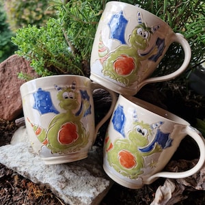 Könnte beinhalten: Drei cremefarbene Keramikbecher, jeder mit einem erhabenen, handbemalten Design eines grünen Drachen mit blauen Flügeln, einem gestreiften Schwanz und einem leuchtend orangefarbenen Bauch. Die Becher haben eine glänzende Oberfläche und einen geschwungenen Henkel.