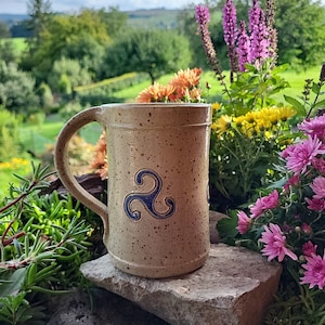 Könnte beinhalten: Eine beige Keramiktasse mit einem blauen und weißen keltischen Knotendesign. Die Tasse steht auf einer Steinoberfläche mit einem verschwommenen Hintergrund aus grünen Bäumen und Blumen.