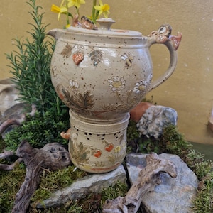Peut inclure: Un ensemble de poterie en céramique en deux parties avec une finition beige mouchetée. La partie supérieure est un pot avec un couvercle et une poignée, décoré de feuilles, de fleurs et d'abeilles. La partie inférieure est un récipient plus petit assorti. Des jonquilles jaunes sont placées dessus.