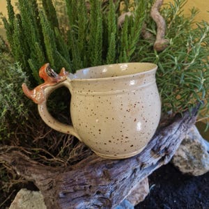 Op de afbeelding: Een gespikkelde keramische mok met een vossenhandvat. De mok is beige met bruine spikkels en een afgeronde vorm. Het vossenhandvat is oranje en bruin. De mok is omgeven door groen en een stuk drijfhout.