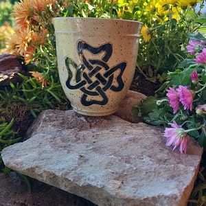 Può includere: Una tazza in ceramica con un designo a nodo celtico nero su uno sfondo beige macchiato. La tazza è appoggiata su una roccia grigio chiaro.
