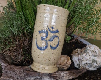 Könnte beinhalten: Beige Keramikvase mit blauem Om-Symbol. Die Vase hat eine gesprenkelte Oberfläche und einen ausgestellten Sockel. Grüne Pflanzen und Steine umgeben die Vase und schaffen eine natürliche Ästhetik.