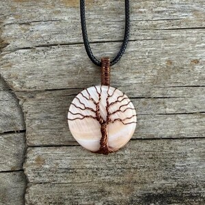 Peut inclure: Un pendentif rond en coquillage blanc irisé orné d'un motif d'arbre de vie en fil de cuivre enroulé sur la surface. Le pendentif est suspendu à un collier en cordon noir, mettant en valeur un tronc métallique détaillé et des branches.
