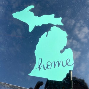 May include: A teal blue outline of the state of Michigan with the word "home" written in cursive inside the state outline.