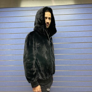 May include: A black fur hooded jacket with a zipper closure. The jacket is worn by a person standing in front of a blue wall.