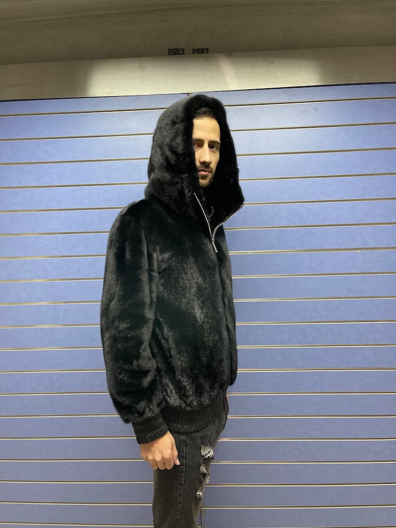 May include: A black fur hooded jacket with a zipper closure. The jacket is worn by a person standing in front of a blue wall.