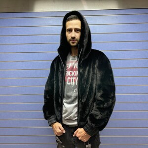 May include: A black faux fur bomber jacket with a zipper closure and a hood. The jacket has a ribbed knit trim on the cuffs and waistband. The model is wearing a grey t-shirt with the text "New York City" printed on it.
