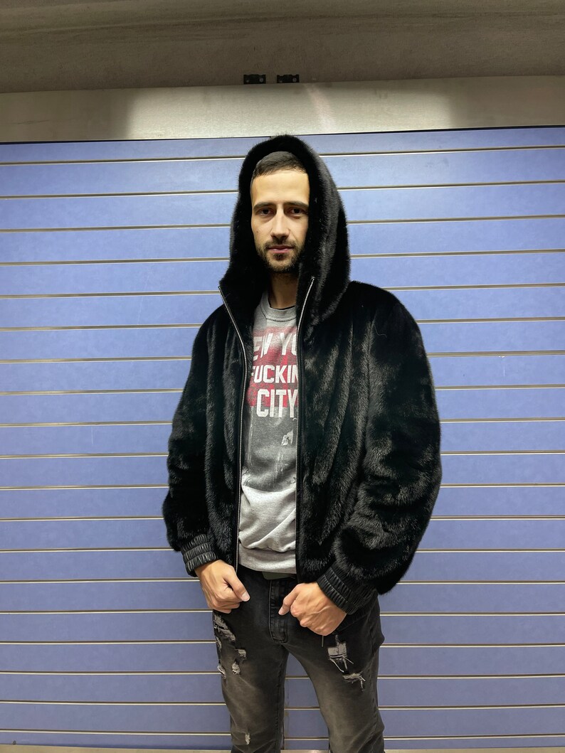 May include: A black faux fur bomber jacket with a zipper closure and a hood. The jacket has a ribbed knit trim on the cuffs and waistband. The model is wearing a grey t-shirt with the text "New York City" printed on it.