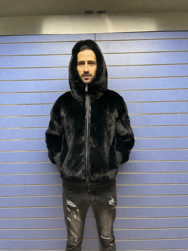 May include: A black fur bomber jacket with a zipper closure and a hood. The jacket is worn by a person standing in front of a blue wall.