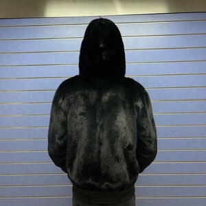 May include: A black fur bomber jacket with a hood. The jacket is worn by a person standing in front of a blue wall.