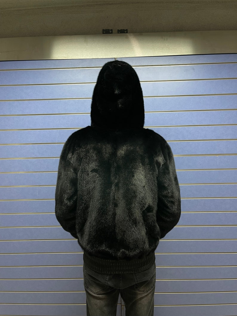May include: A black fur bomber jacket with a hood. The jacket is worn by a person standing in front of a blue wall.