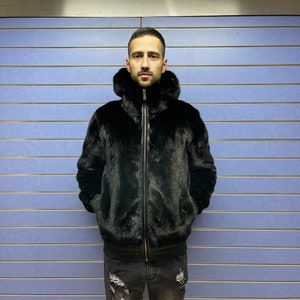 May include: A black fur bomber jacket with a zipper closure and a hood. The jacket is worn by a person standing in front of a blue wall.