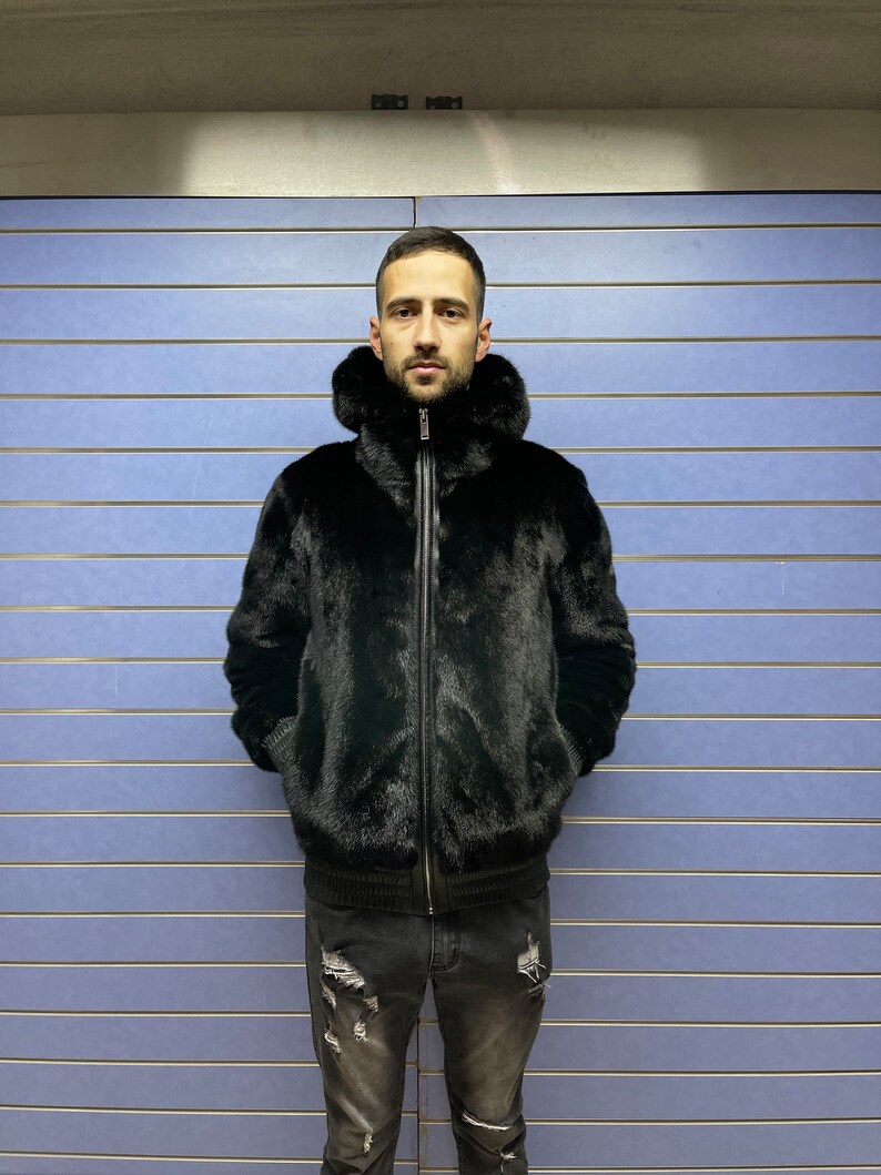 May include: A black fur bomber jacket with a zipper closure and a hood. The jacket is worn by a person standing in front of a blue wall.