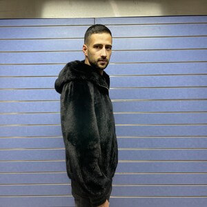 May include: A black fur jacket with a hood. The jacket is zipped up and the person is wearing black pants.