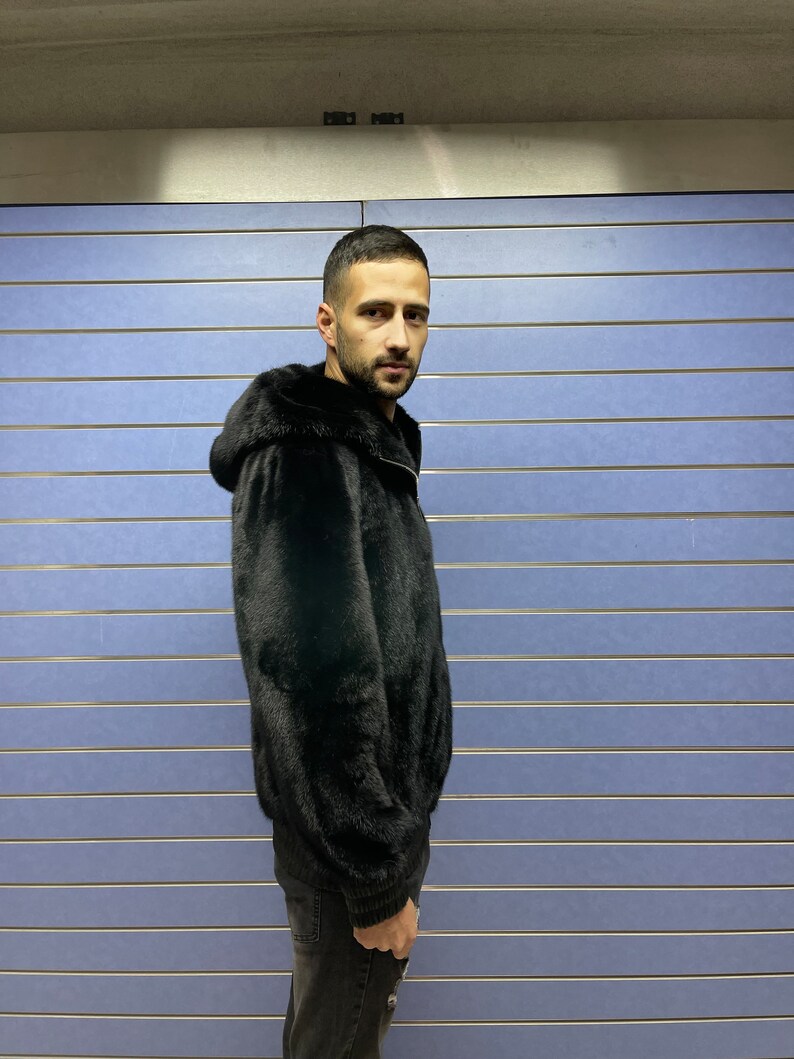 May include: A black fur jacket with a hood. The jacket is zipped up and the person is wearing black pants.