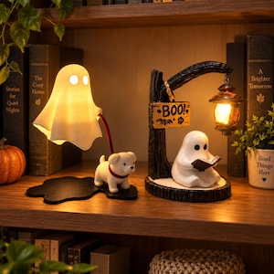 May include: Two decorative ghost-themed lamps. One lamp features a white ghost sheet over a light with a small white dog on a black base. The second lamp shows a white ghost reading a book under a hanging lantern with a sign that reads BOO.