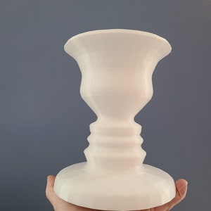 May include: White ceramic vase with a face illusion design. The vase is shaped like a traditional urn with a wide base and a narrow neck.