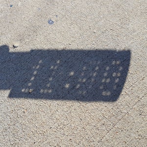 Puede incluir: La sombra de una farola proyectada sobre una superficie de hormigón. La sombra muestra la forma de la farola con un patrón de rejilla.