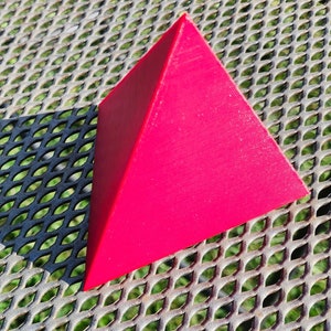 May include: A bright red pyramid-shaped object made of plastic. The object is sitting on a metal surface with a grid pattern.