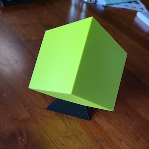 May include: A bright green cube shaped object sits on a black triangular stand. The cube is made of a smooth, glossy material.