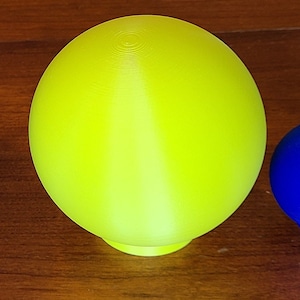May include: A bright yellow sphere with a smooth, slightly textured surface. The sphere is sitting on a wooden surface.