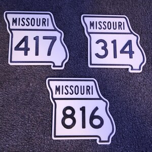 May include: Three white, state-shaped signs with black borders and lettering. Each sign features the word MISSOURI at the top and a large area code number in the center. The numbers displayed are 417, 314, and 816, all in a bold, black font.