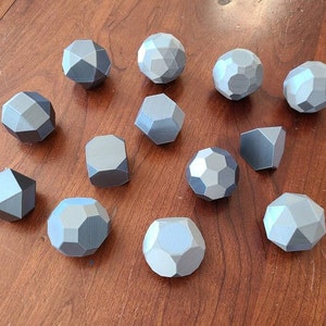 May include: A selection of geometric shapes, including spheres and polyhedrons, in shades of grey. The objects are arranged on a wooden surface. The shapes are 3D printed and have a modern, minimalist aesthetic. Measurements are in centimeters.