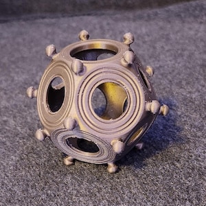 May include: A metallic-coloured, geometric object with a spherical shape. It features circular openings and protruding rounded elements. The object appears 3D-printed and sits on a textured surface.