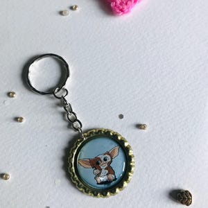 May include: A keychain featuring a cartoon Gremlin character inside a bottle cap. The keychain has a silver ring and chain. A pink crocheted heart and small gold beads are scattered around the keychain on a white surface.