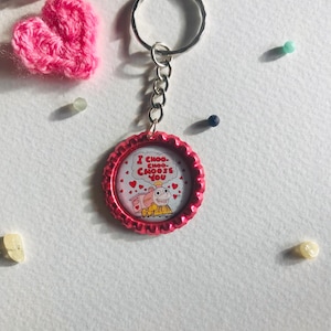 May include: A pink bottle cap keychain with a silver keyring and chain. The bottle cap features a cartoon train with the text "I CHOO-CHOO-CHOOSE YOU" surrounded by hearts. A pink crocheted heart and small beads are also visible.