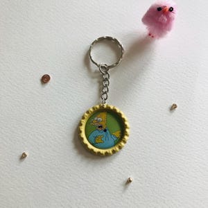 Op de afbeelding: Gele en groene sleutelhanger met een cartoon afbeelding van een geel personage met een blauw shirt en een verraste uitdrukking. De sleutelhanger is bevestigd aan een zilveren sleutelring.