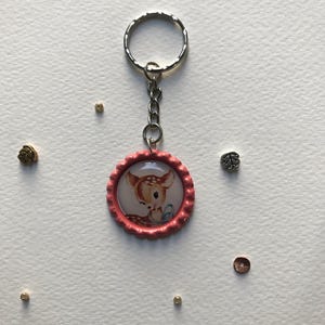 Op de afbeelding: Een sleutelhanger met een roze flesdopontwerp met een hertenillustratie. De sleutelhanger heeft een zilveren ring en ketting. De hertenillustratie staat in een cirkelvormige lijst. De achtergrond is wit.