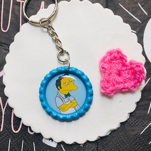 Könnte beinhalten: Ein Schlüsselanhänger mit einem Cartoon-Bild von Moe Szyslak von den Simpsons, in einem blauen Flaschenverschluss. Der Schlüsselanhänger hat eine silberne Kette und einen Ring. Ein kleines, rosa gehäkeltes Herz befindet sich neben dem Schlüsselanhänger.